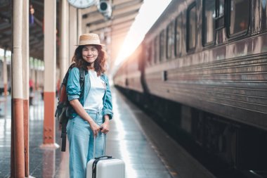Sırt çantalı ve bavullu gülümseyen genç bir kadın tren istasyonunda bekliyor, seyahat etmeye hazır..