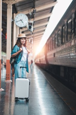 Elinde bavul ve sırt çantası olan genç bir kadın tren istasyonu peronunda yolculuk için hazır bekliyor. Güneş arka planda doğar..