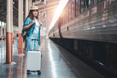 Tren istasyonunda bavulu ve sırt çantasıyla gülümseyen kadın, seyahat macerasına hazır..