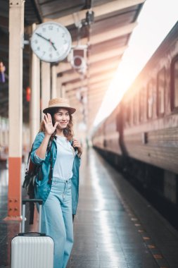 Tren istasyonunda günlük kıyafet dalgaları içinde gülümseyen kadın, seyahat macerasına hazır. Güneş ışığı platformu aydınlatır.