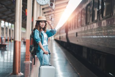 Sıradan giyinmiş ve şapkalı genç bir kadın tren istasyonunda bir bavulla gezmeye hazır bekliyor..