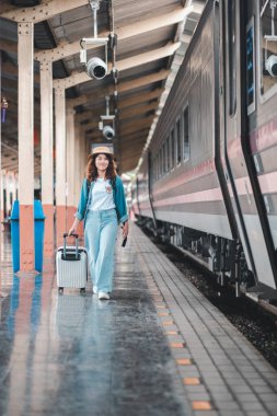 Elinde bavuluyla gülümseyen kadın tren istasyonunda yürüyor, yolculuğa ve maceraya hazır..