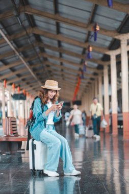 * * Genç bir kadın tren istasyonunda, akıllı telefonunu kullanarak bavulunun üzerine oturur. Hasır şapka takıyor ve günlük giysiler giyiyor. Seyahate hazır.