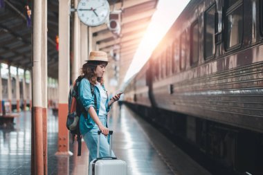 Günlük kıyafetleri ve şapkası olan genç bir kadın tren istasyonunda elinde bir bavulla güneş ışığı içeri girerken akıllı telefonunu kontrol ediyor..