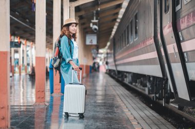 Elinde bavul olan genç bir kadın tren istasyonunda duruyor, yolculuğa hazır. Macera ruhunu somutlaştıran günlük giysiler ve bir şapka giyiyor..