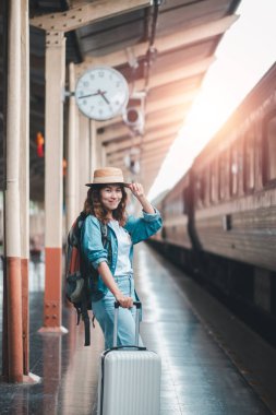 Tren istasyonunda çantalı ve sırt çantalı gülümseyen genç bir kadın, seyahat ve maceraya hazır..
