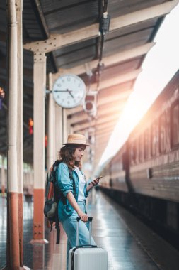 Bavullu bir kadın yolcu tren istasyonunda akıllı telefon kullanıyor, yolculuk için hazır. Platformdan gün ışığı akıyor.
