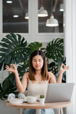 Genç bir kadının, yemyeşil bitkiler ve yumuşak doğal ışıkla çevrili bir dizüstü bilgisayarla meditasyon yaptığı huzurlu bir sahne..