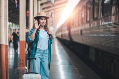 Genç bir kadın, elinde bir bavulla tren istasyonunda el sallıyor. Platformdan güneş ışığı geçiyor..