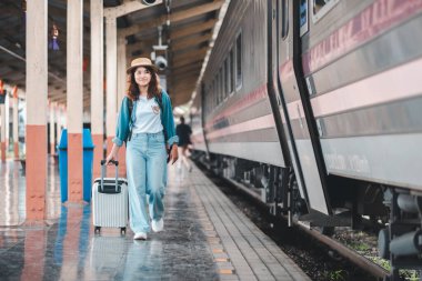 Sıradan giyinmiş ve şapkalı genç bir kadın tren istasyonunda bir bavulla yürüyor, seyahat ve maceranın vücut bulmuş hali..