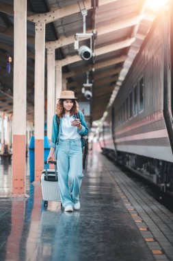 Gündelik giyinmiş ve şapkalı genç bir kadın, elinde bavuluyla, güneşli bir platformda tren beklerken telefonunu kontrol ediyor..