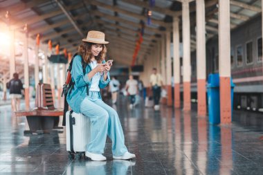 Yaz kıyafetleri içinde genç bir kadın tren istasyonunda bir bavula oturur, akıllı telefonunu kullanarak, seyahat maceralarına hazırlanır..