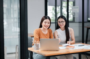 Modern bir ofiste birlikte çalışan iki genç kadın, fikirlerini dizüstü bilgisayarla ve masada belgelerle tartışıyor..