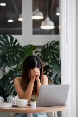 Modern ofis ortamında yemyeşil bitkilerle dolu bir masada dizüstü bilgisayarla oturan bir kadın stresli görünüyor..