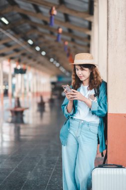 Tren istasyonundaki kadın gezgin, akıllı telefon kullanıyor, günlük kıyafetler ve hasır şapka giyiyor, bavullu..