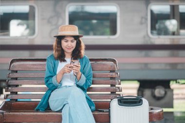 Günlük kıyafetli ve hasır şapkalı genç bir kadın tren istasyonunda akıllı telefon kullanıyor, bankta bavullarıyla oturuyor..