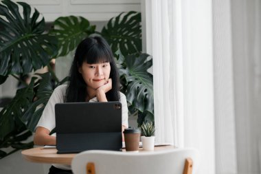 Genç bir kadın, yemyeşil bitkiler ve yumuşak doğal ışıkla çevrili, rahat bir ofis ortamında tabletine odaklanmış..