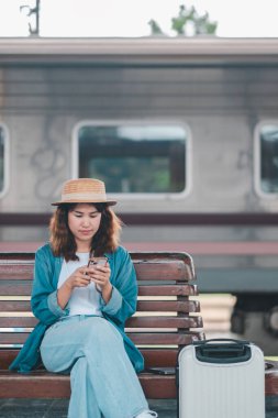 Şapkalı ve kot ceketli bir kadın tren istasyonunda bavuluyla oturmuş, akıllı telefon kullanıyor, treni bekliyor..