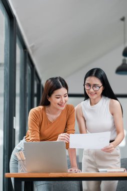 Parlak bir ofiste birlikte çalışan iki genç kadın, belgeleri gözden geçiriyorlar ve dizüstü bilgisayar kullanıyorlar..