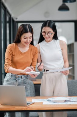 Parlak bir ofiste birlikte çalışan iki genç kadın, belgeleri tartışıyor ve dizüstü bilgisayar kullanıyorlar..