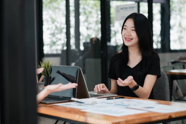 Modern ofis ortamında genç bir kadın, bir meslektaşıyla iş görüşmesi yapıyor, dizüstü bilgisayar kullanıyor ve belgeleri inceliyor..