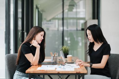 Modern bir ofiste birlikte çalışan iki kadın, iş planlarını dizüstü bilgisayarlarla ve ahşap bir masadaki belgelerle tartışıyorlar..