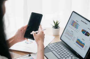 Parlak ofis ortamında finansal çizelge ve grafikleri incelemek için akıllı telefon ve dizüstü bilgisayar kullanan profesyonel bir kadın..