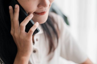 Beyaz tişörtlü bir kadın kulağına akıllı bir telefon dayar, sohbet eder, arka planı bulanıktır..