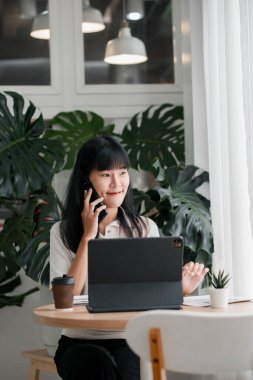 * * Genç bir kadın, yemyeşil bitkilerle çevrili şık bir ofis ortamında, tablet ve telefonla birden fazla iş yapıyor.