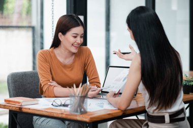 Modern ofis ortamında dizüstü bilgisayarlarla ve çizelgelerle iş stratejilerini tartışan, takım çalışması ve işbirliği sergileyen iki kadın..