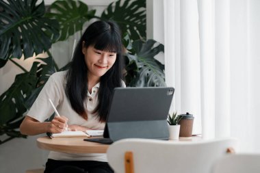 Genç bir kadın elinde dizüstü bilgisayar, defter ve kahveyle büyük yeşil bitkilerle dolu parlak bir odada oturuyor..