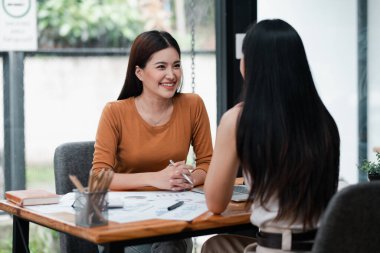 İki kadın bir masada oturuyor, gülümsüyor ve modern ofis ortamında dökümanlar ve kahve fincanlarıyla konuşuyor..