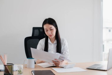 Modern ofis ortamında, masasında doğal ışıkla dokümanları incelemeye odaklanmış genç bir kadın..