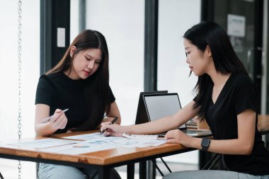 Masada birlikte çalışan iki kadın, belgeleri gözden geçiriyorlar ve modern ofis ortamında dizüstü bilgisayar kullanıyorlar..