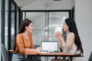 Modern ofis ortamında masa başında sohbet eden iki kadın, dizüstü bilgisayar ve kırtasiye malzemesi, takım çalışması ve işbirliği.