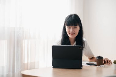 Evde bir ofis ortamında tabletine odaklanmış genç bir kadın, yanında kahve ve belgelerle, doğal ışıkla aydınlatılmış..