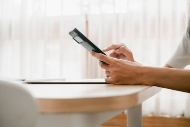 Parlak bir odada, perdeleri ve doğal ışığı olan ahşap bir masada akıllı telefon kullanan ellerin yakın çekimi..