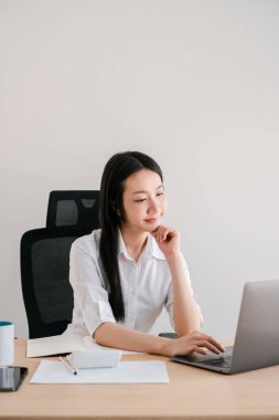 Beyaz gömlekli genç bir kadın parlak, minimalist ofis ortamında bir masada dizüstü bilgisayarla çalışıyor..