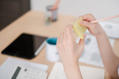 Tablet, kupa ve hesap makinesi gibi ofis malzemeleriyle çevrili, masanın üzerinde bir kalemle yapışkan bir not tutan ellerin yakın çekimi..