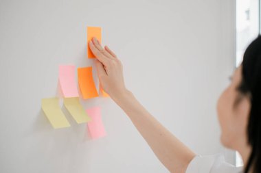 Bir kadın beyaz bir duvara renkli yapışkan notlar yerleştiriyor. Modern bir çalışma alanındaki yaratıcı planlama ve organizasyonu simgeliyor..