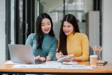 İki kadın bir masada dizüstü bilgisayar ve defterlerle birlikte çalışıyor, modern ofis ortamında takım çalışması ve işbirliği sergiliyorlar..