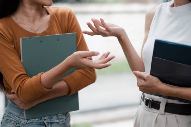 Two women discussing ideas while holding notebooks, showcasing communication and collaboration in an informal environment. clipart