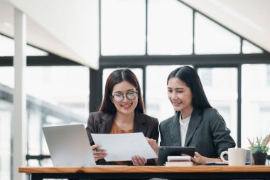 Parlak, modern bir ofiste birlikte çalışan iki profesyonel kadın, belgeleri gözden geçiriyorlar ve bir dizüstü bilgisayar kullanıyorlar..