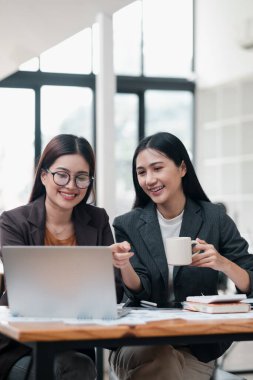 İş kıyafeti giymiş iki kadın, parlak ve modern ofis ortamında dizüstü bilgisayarda birlikte çalışıyorlar..