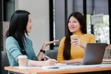 Modern ofis ortamında gülümseyen ve sohbet eden iki genç kadın, masada dizüstü bilgisayarları ve defterleri var..