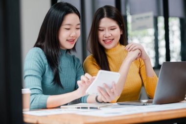 Modern ofis ortamında, akıllı telefon ve dizüstü bilgisayar kullanan iki genç kadın, ahşap bir masanın üzerinde belgeleri olan bir projede işbirliği yapıyorlar..