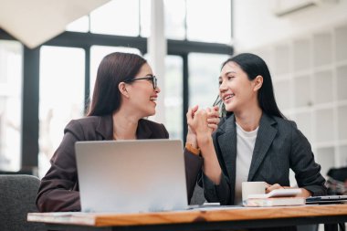 Modern bir ofiste iki iş kadını, gülümsüyor ve bir masada dizüstü bilgisayar ve defterlerle birlikte çalışıyorlar..
