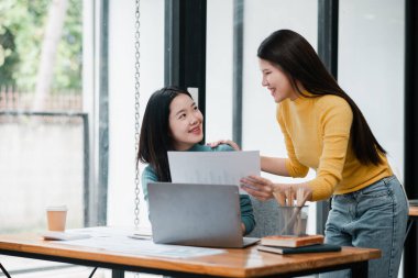 Modern bir ofiste birlikte çalışan iki genç kadın, belgeleri tartışıyor ve dizüstü bilgisayar kullanıyor, işbirlikçi ve pozitif bir çalışma ortamı yaratıyor..