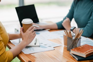 İki meslektaş modern bir ofiste işbirliği yapıyor, kahve eşliğinde belgeler ve fikirleri gözden geçiriyorlar, masada dizüstü bilgisayarlar ve kırtasiyeler var..