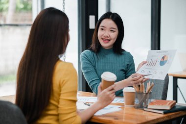 Modern bir ofiste iki kadın satış analizini tartışıyor, ellerinde kahve fincanları ve çizelgeleri var..